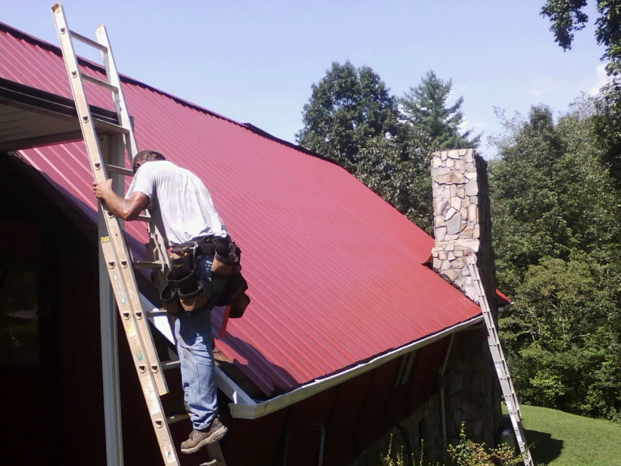 Roofer on a ladder beside a red metal roof and stone chimney — Soffit & Fascia Repair by Roofing Brothers in Iron Ridge
